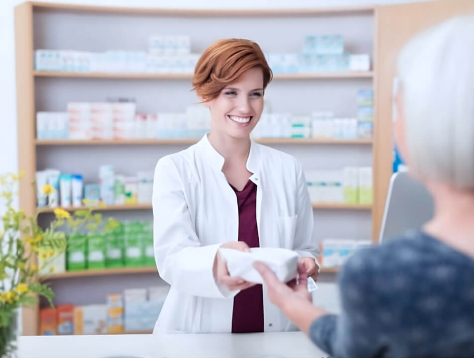 Eine Frau mit weißem Kittel steht in der Offizin einer Apotheke und übergibt einer anderen Frau ein Medikament.