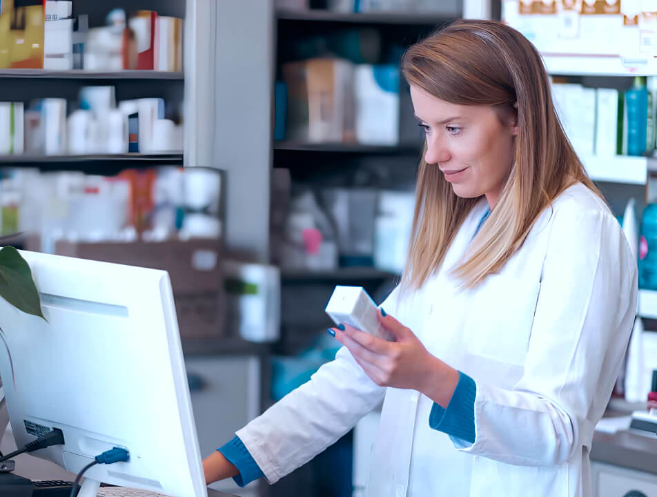 Eine Frau steht in einer Offizin, hält ein Medikament in der Hand und schaut gleichzeitig auf den Computer.
